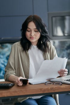 Young woman sitting indoors, reviewing bills and using smartphone.
